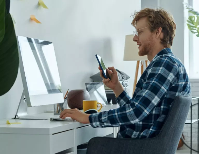 A man in a plaid shirt sits at a desk, looking at his smartphone and working on a computer.