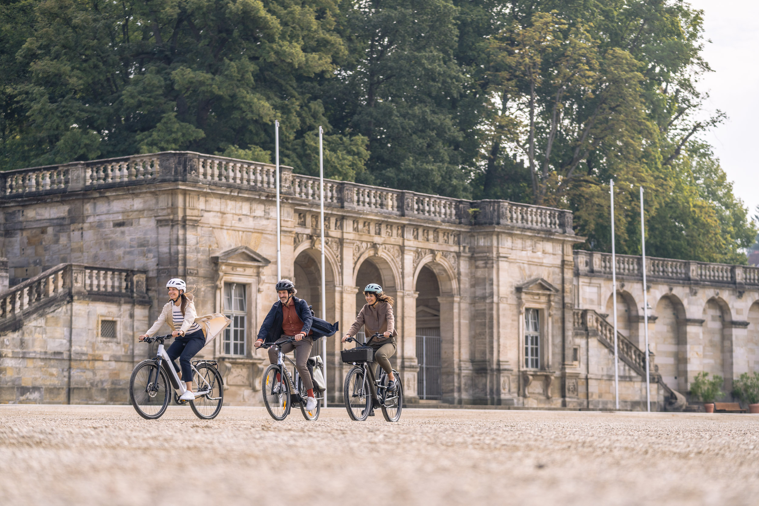 Drei Personen fahren mit E-Bikes vor einem historischen Gebäude mit Steinarkaden entlang.