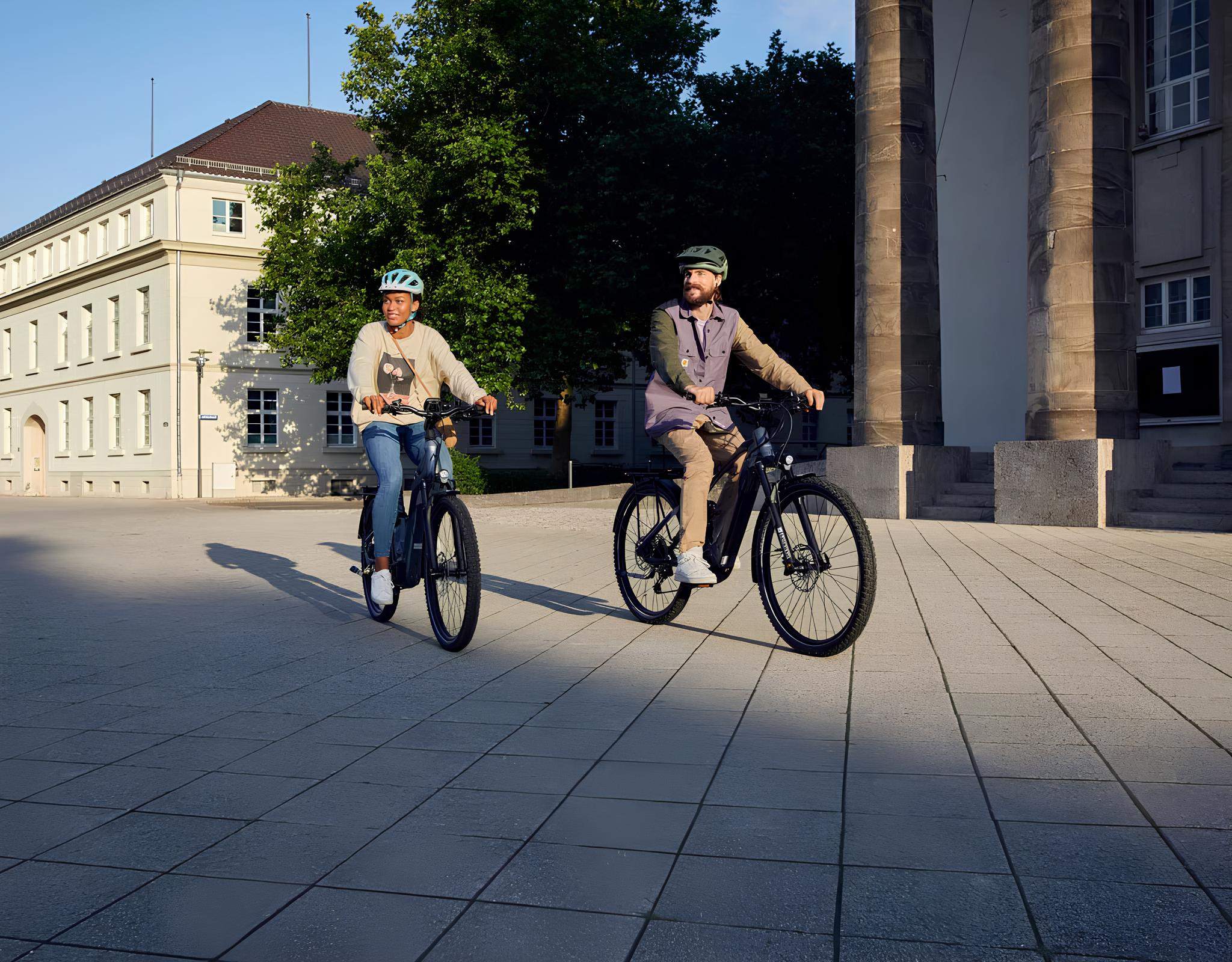 Two people are riding e-bikes on a paved square in front of a historic building in sunny weather.