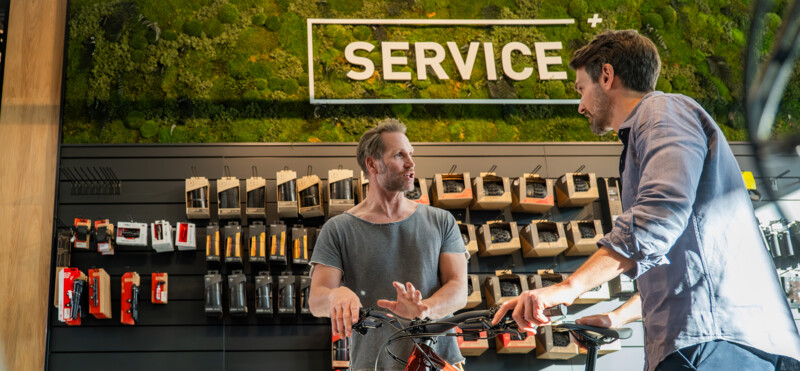 Two men are talking in a bicycle repair shop in front of a service wall while discussing a bike.