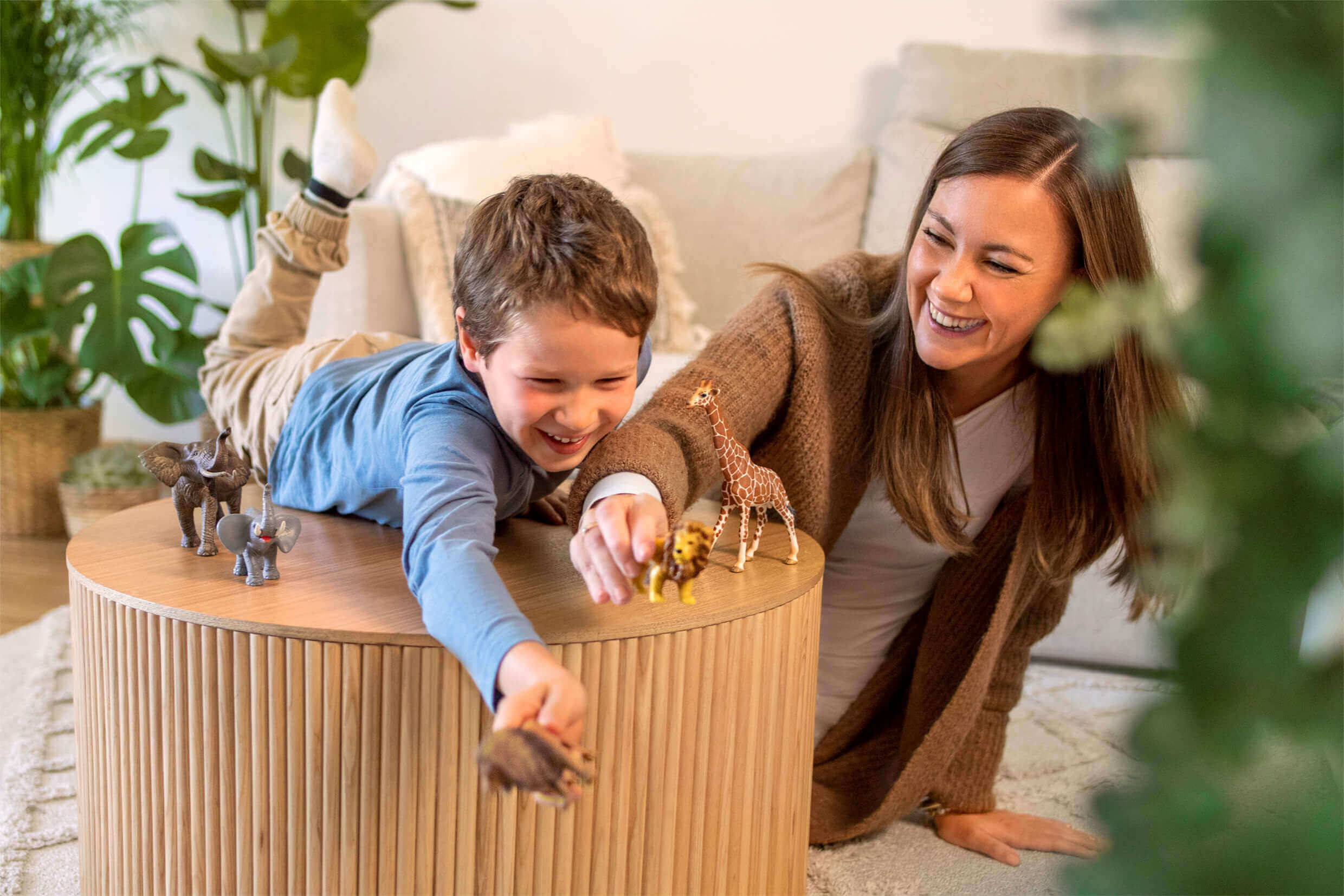 A smiling woman plays with a little boy lying on a round wooden table. Both are moving small toy animals such as elephants, lions, and giraffes. Green houseplants and a sofa can be seen in the background.