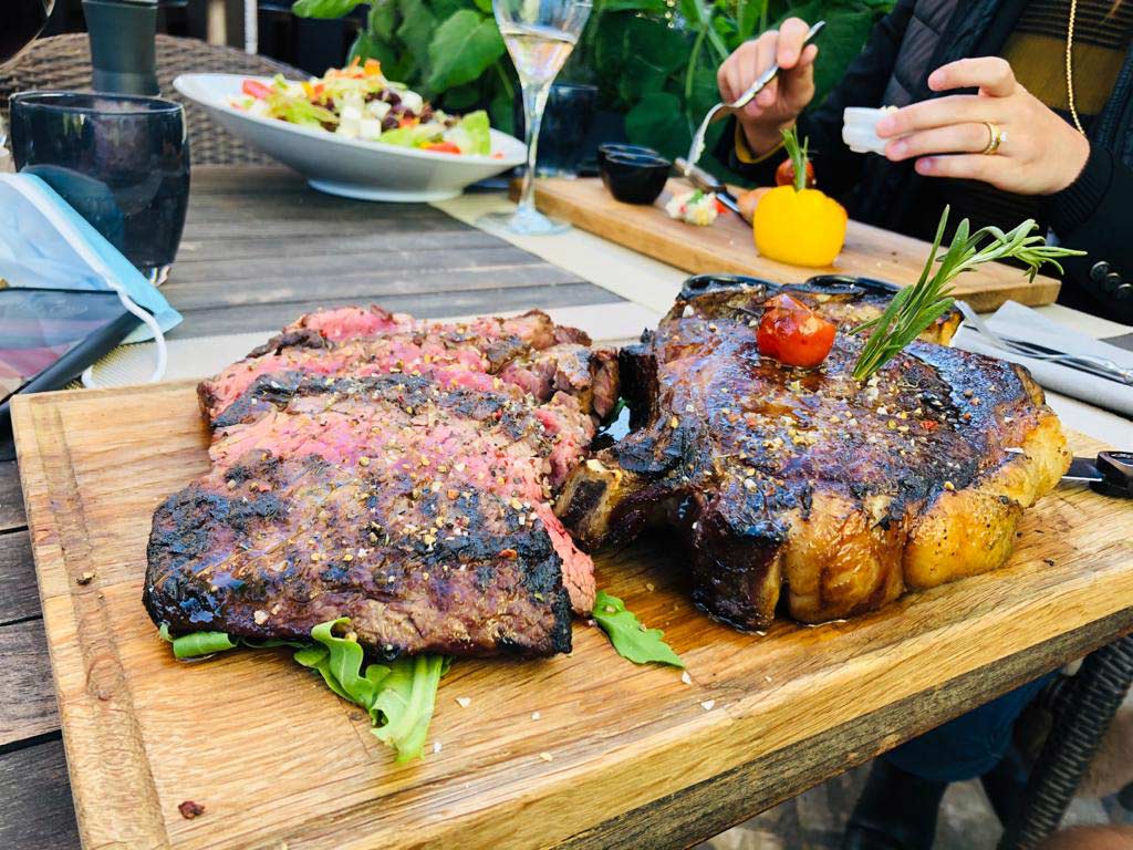 Two grilled steaks, one sliced, garnished with rosemary on a wooden board. In the background, a salad and a person eating.