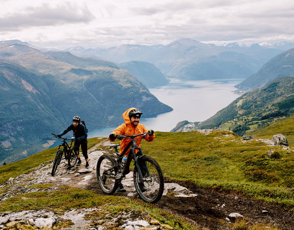 Two mountain bikers push their bikes up a steep mountain trail, with a sweeping view of a lake and surrounding mountains in the background.