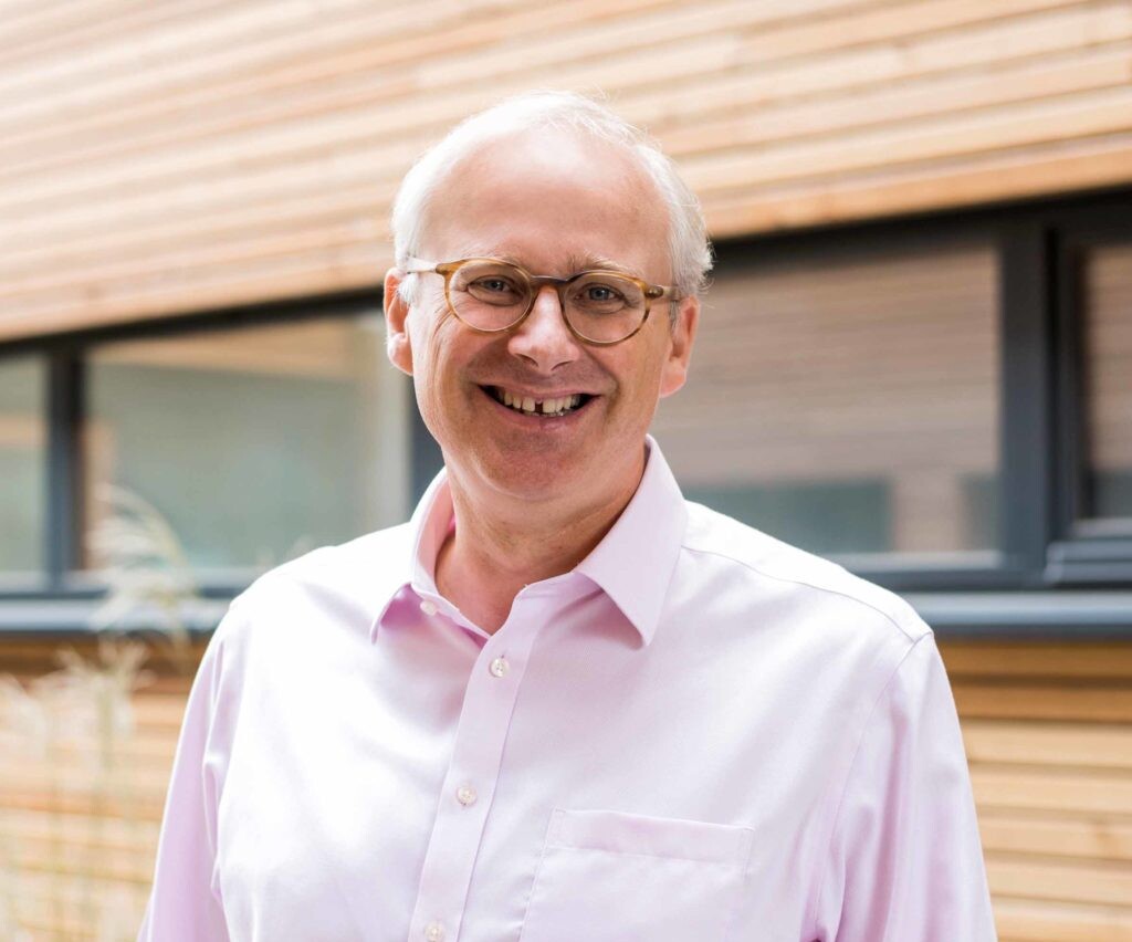 Portrait of a man wearing glasses and a pink shirt, smiling in front of a building with wood paneling.